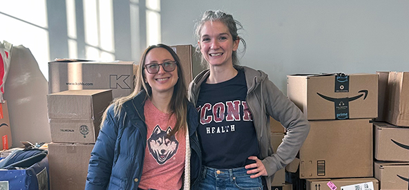 Two ambulatory social workers, Rachel Boxwell and Eleanor Szmurlo, standing in front of Amazon boxes for a holiday toy drive.